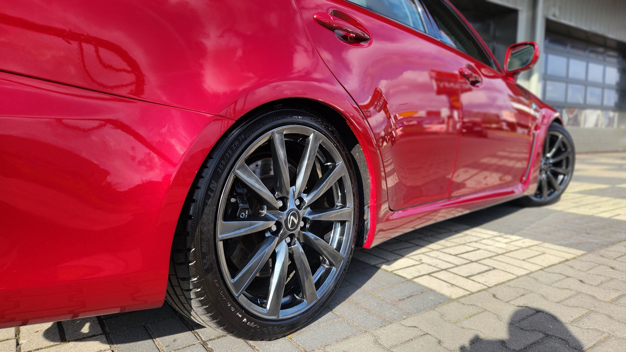 Błyszczący, czerwony Lexus IS z polakierowanymi felgami, zaparkowany na kostce brukowej w słoneczny dzień, widok z boku.