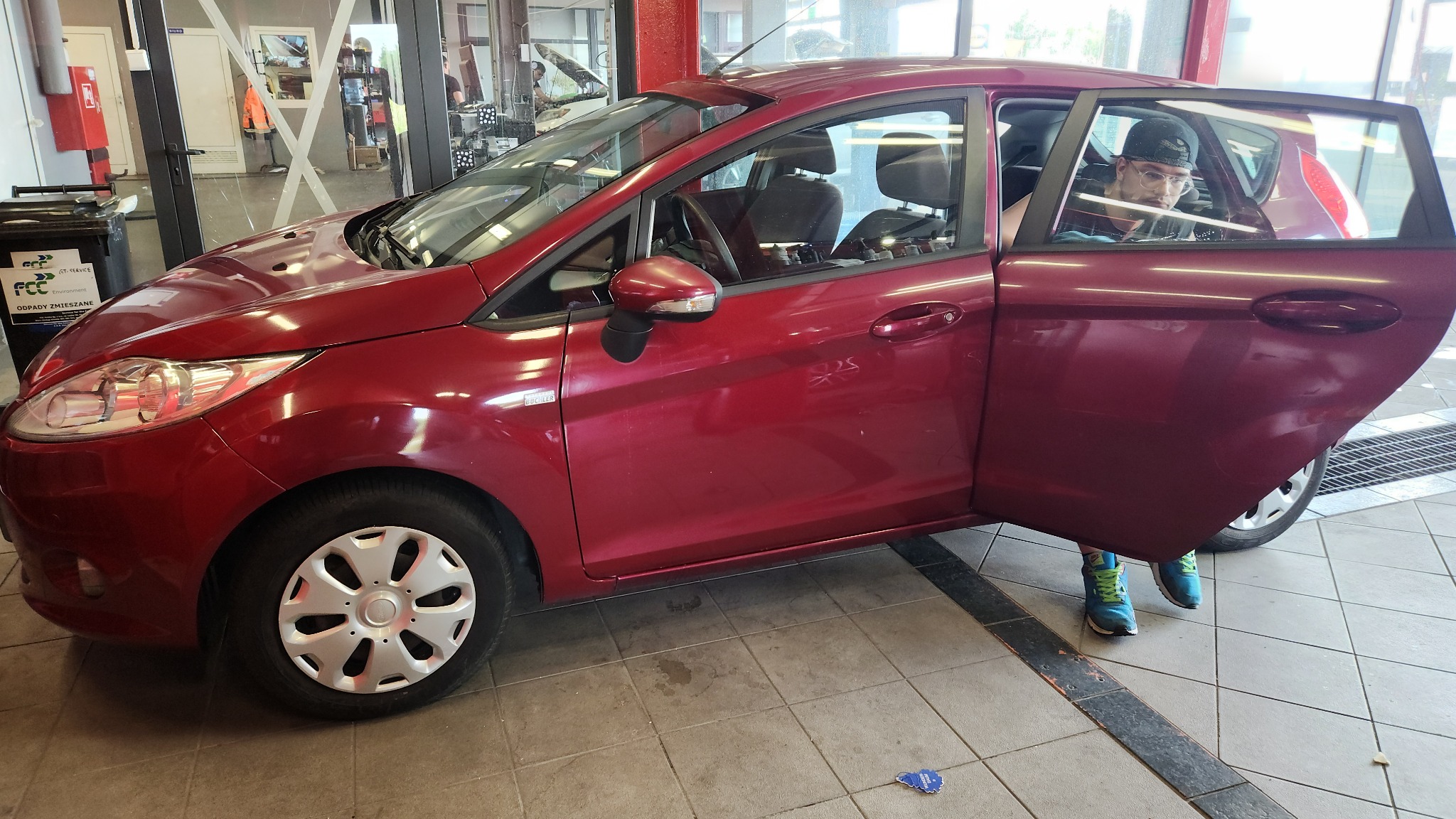 Czerwony Ford Fiesta z otwartymi drzwiami pasażera, wewnątrz widoczny mężczyzna w czapce i okularach, stojący na tle wnętrza myjni samochodowej.