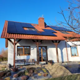 Panel słoneczny zamontowany na dachu domu jednorodzinnego z czerwoną dachówką, widok z poziomu ogrodu w słoneczny dzień.