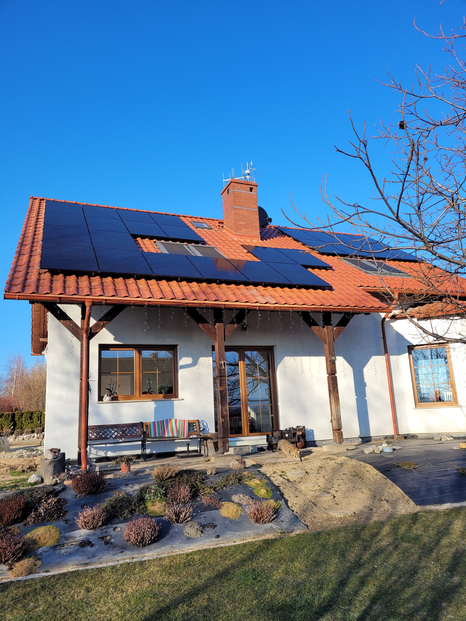 Panel słoneczny zamontowany na dachu domu jednorodzinnego z czerwoną dachówką, widok z poziomu ogrodu w słoneczny dzień.