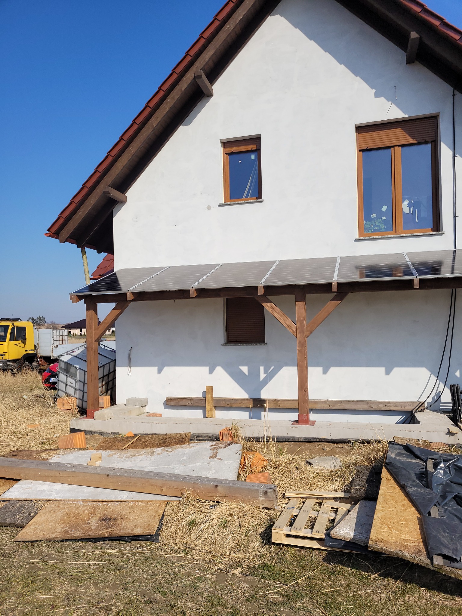Biały dom z drewnianą pergolą, na której zamontowane są panele, prawdopodobnie fotowoltaiczne, widoczne materiały budowlane i żółta ciężarówka w tle.