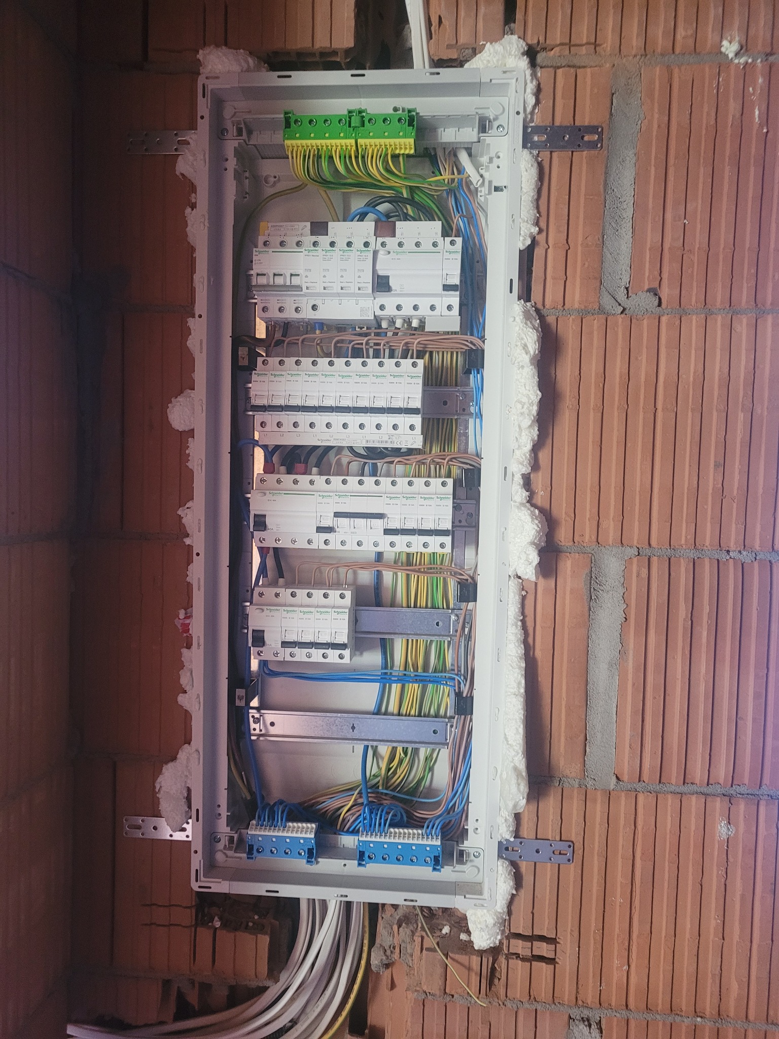 Rozdzielnica elektryczna zamontowana w ścianie z cegły, uszczelniona pianką montażową, z widocznymi przewodami i bezpiecznikami.