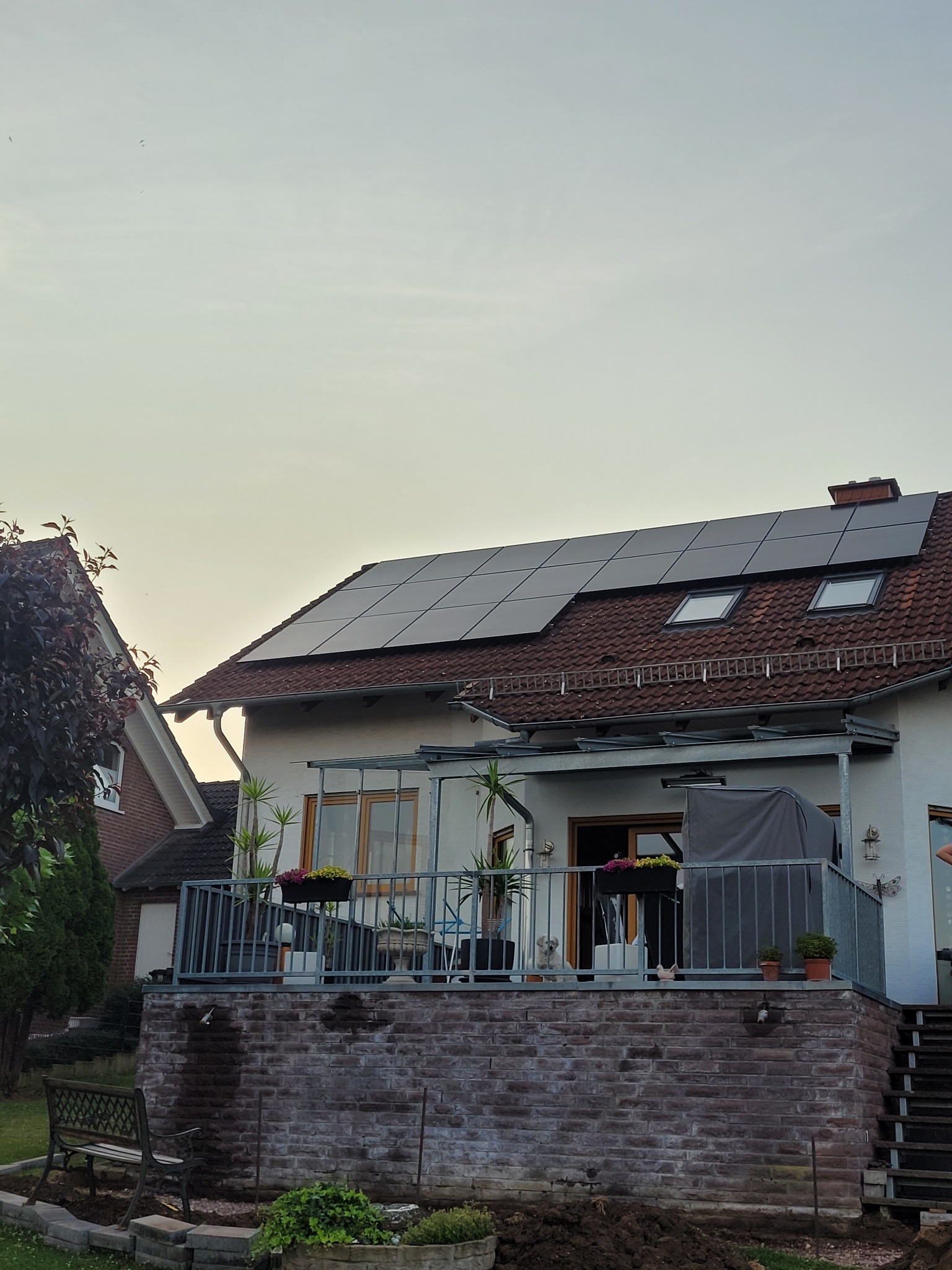 Dom jednorodzinny z panelami fotowoltaicznymi na dachu i balkonem z kwiatami, widok z poziomu ogrodu.