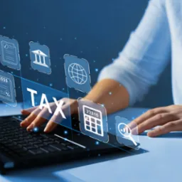 Kobieta w błękitnej koszuli wpisuje dane na laptopie; nad klawiaturą unoszą się ikony związane z finansami, takie jak kalkulator, wykresy, dokumenty i napis TAX.