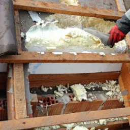 Uszkodzona piana izolacyjna na poddaszu, widoczne drewniane elementy konstrukcyjne, fragment dachu z dachówką, pracownik w czerwonej rękawicy podczas naprawy izolacji.