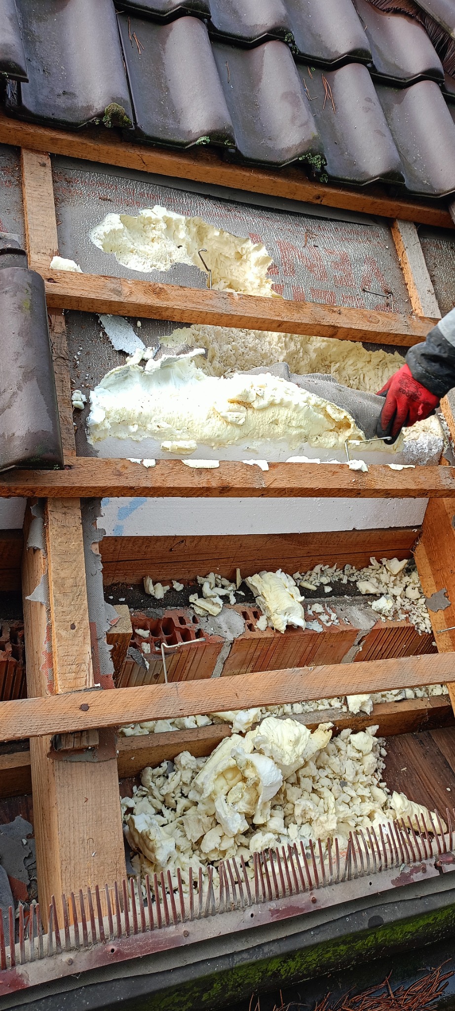 Uszkodzona piana izolacyjna na poddaszu, widoczne drewniane elementy konstrukcyjne, fragment dachu z dachówką, pracownik w czerwonej rękawicy podczas naprawy izolacji.