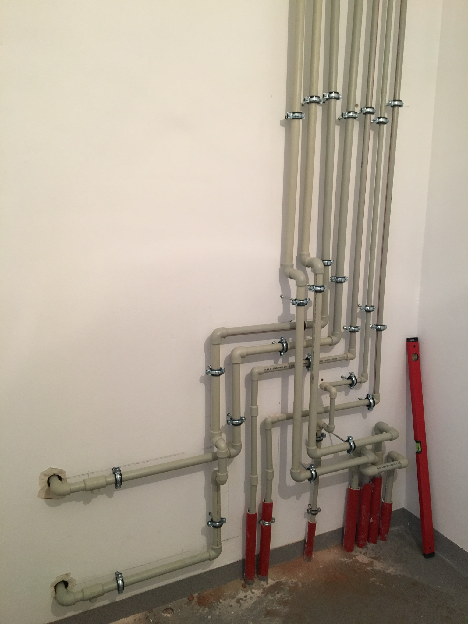 Szare rury instalacji hydraulicznej zamocowane do ściany za pomocą metalowych obejm, z czerwonymi fragmentami u dołu, obok czerwona poziomica oparta o ścianę.