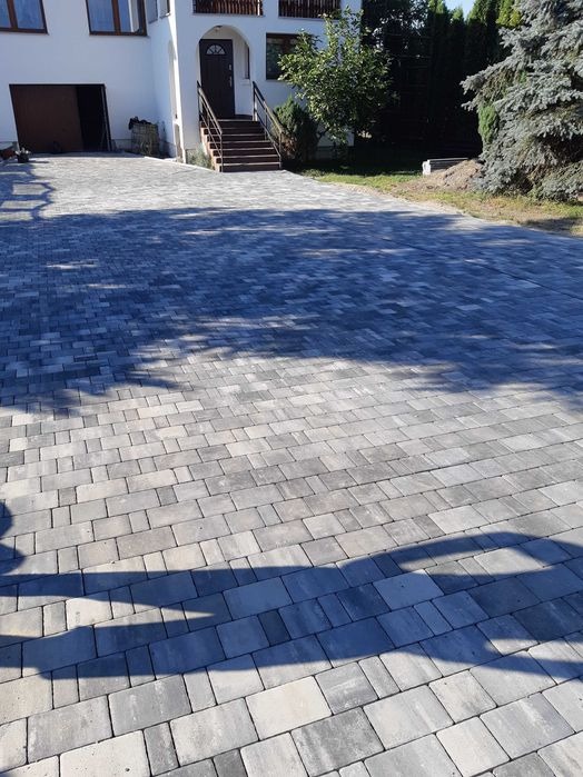 Nowo ułożony podjazd z szarej kostki brukowej przed białym domem z brązowymi schodami i drzwiami.