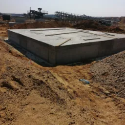 Wylany, betonowy fundament o regularnym kształcie na placu budowy, z wystającymi prętami zbrojeniowymi, otoczony piaskiem i żwirem, z budowlanymi maszynami i konstrukcjami w tle pod błękitnym niebem.