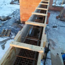 Szczegółowy widok z góry na szalunek z desek i zbrojenie stalowe wewnątrz, przygotowane do zalania betonem, z częściowo wzniesionym murem z czerwonej cegły dziurawki w tle, na placu budowy pokrytym...