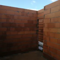 Wnętrze budynku w trakcie budowy z czerwonej cegły ceramicznej, widoczne fragmenty ścian, otwór drzwiowy i rolki materiału izolacyjnego w tle.
