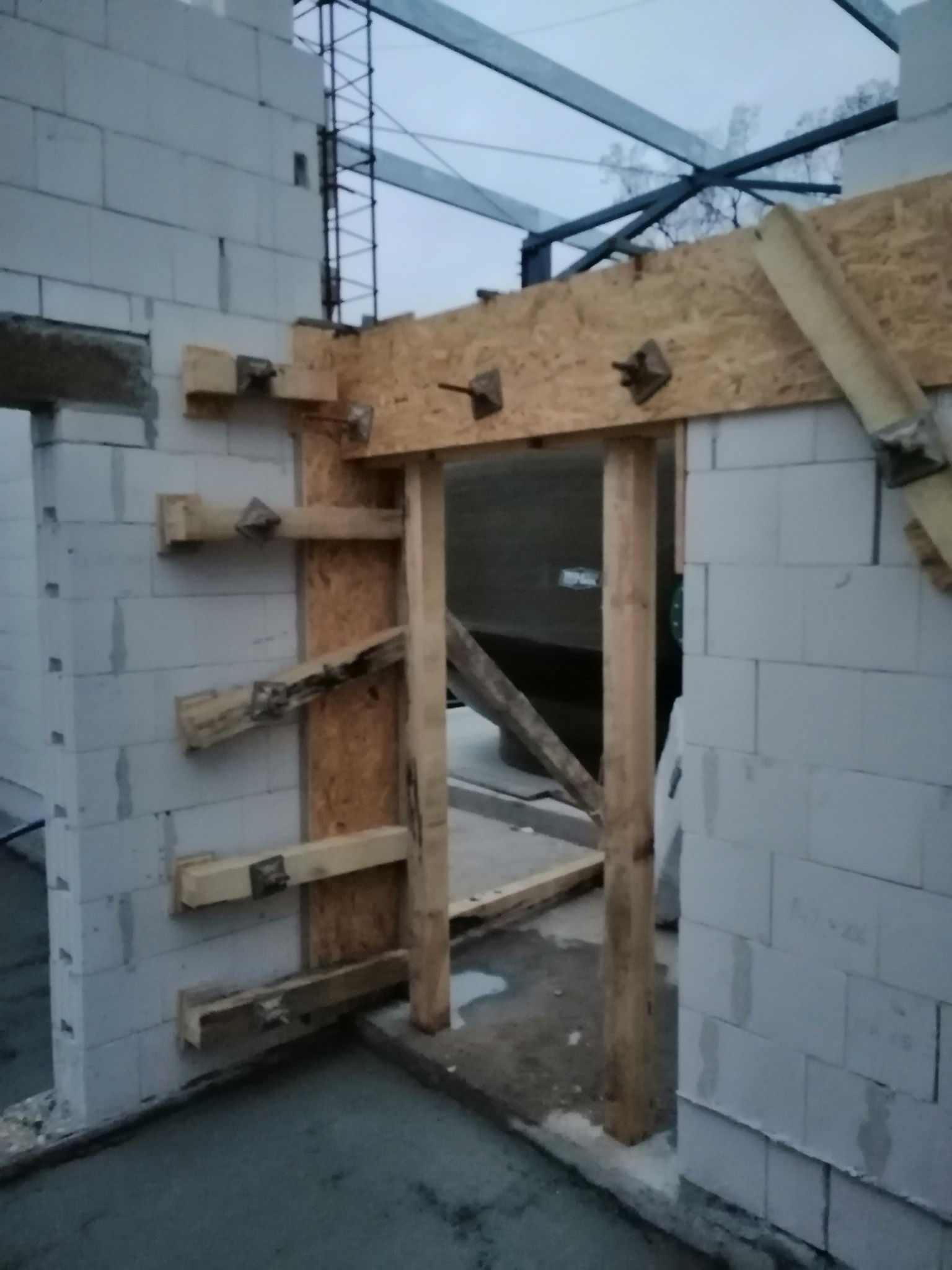 Wznoszenie ściany z betonu komórkowego z drewnianą konstrukcją szalunkową otworu drzwiowego, widoczne drewniane podpory i metalowe łączniki.