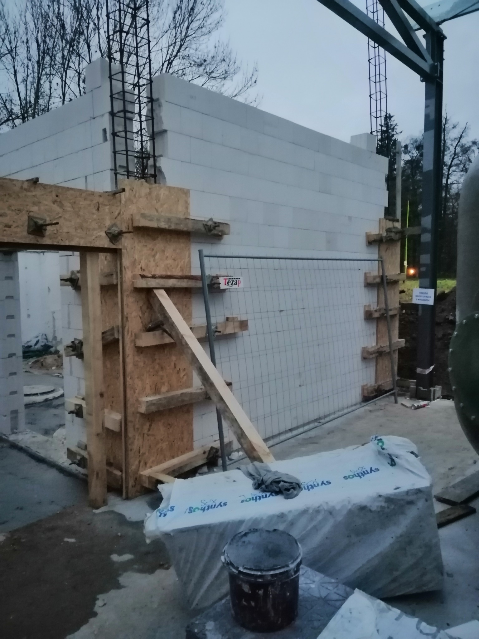 Wznoszenie ścian z białych bloczków, widoczne zbrojenie pionowe, szalunki drewniane przy otworze drzwiowym, teren budowy w pochmurny dzień.