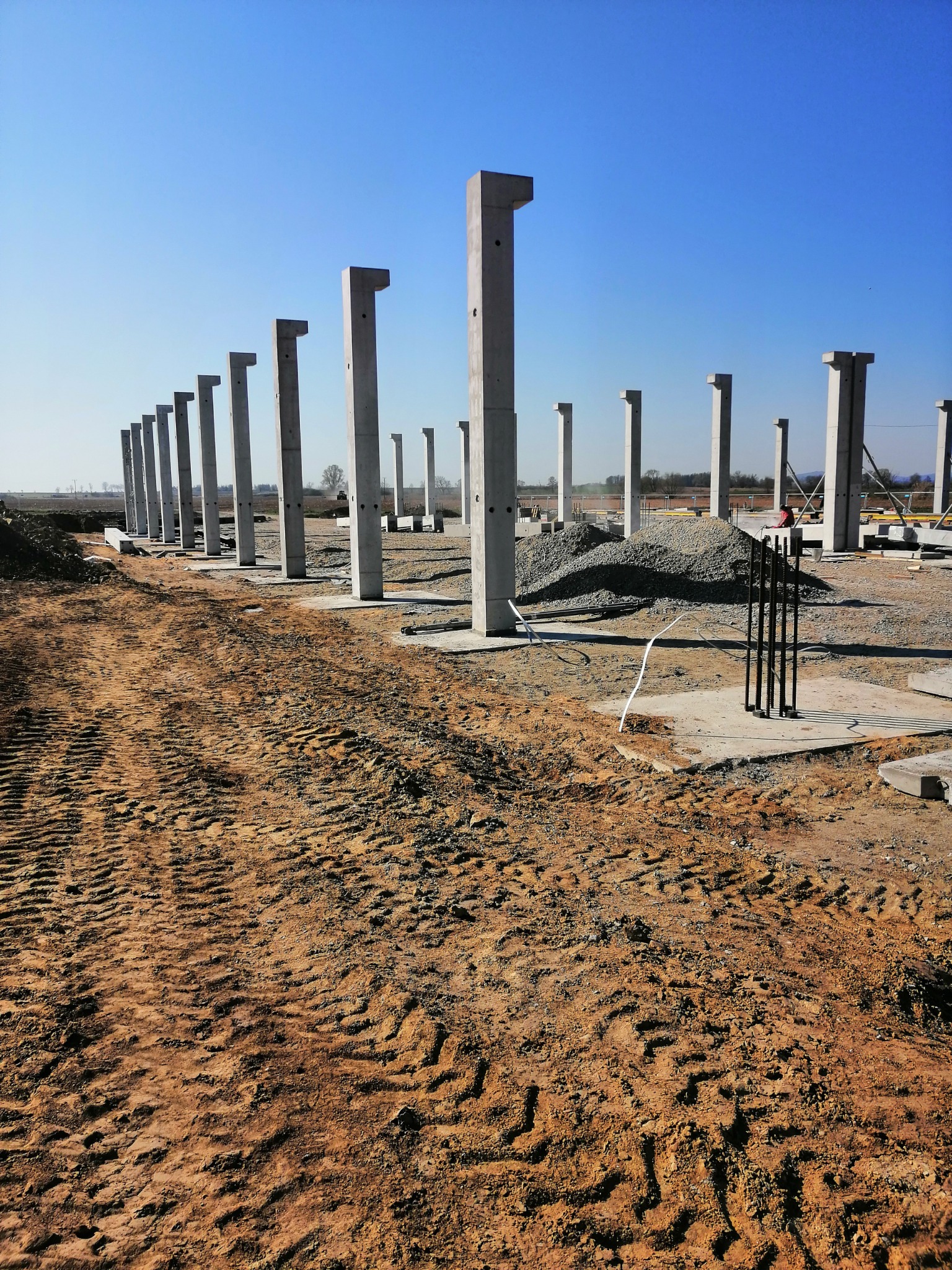 Wznoszenie szkieletu budynku z prefabrykowanych słupów żelbetowych na placu budowy, widoczne ślady opon ciężkiego sprzętu na ziemi i hałda kruszywa w tle.