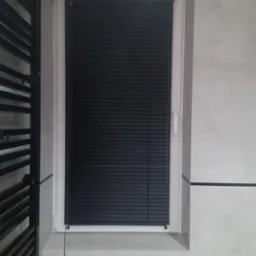 Białe okno z czarnymi żaluzjami wewnętrznymi w łazience z szarą ścianą i czarnym grzejnikiem drabinkowym po lewej stronie.