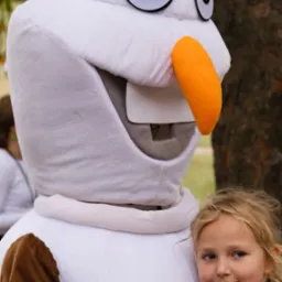 Dziewczynka przytulona do pluszowego kostiumu bałwana Olafa z 'Krainy Lodu', widok z bliska, zarys drzew w tle.