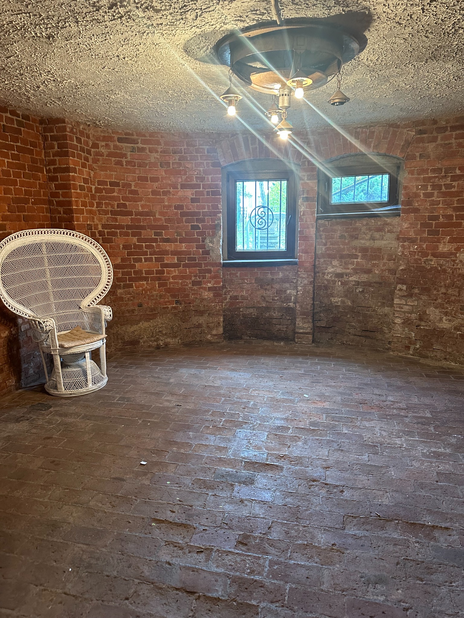 Wnętrze piwnicy z ceglanymi ścianami, oświetlone wiszącą lampą, z białym wiklinowym fotelem.