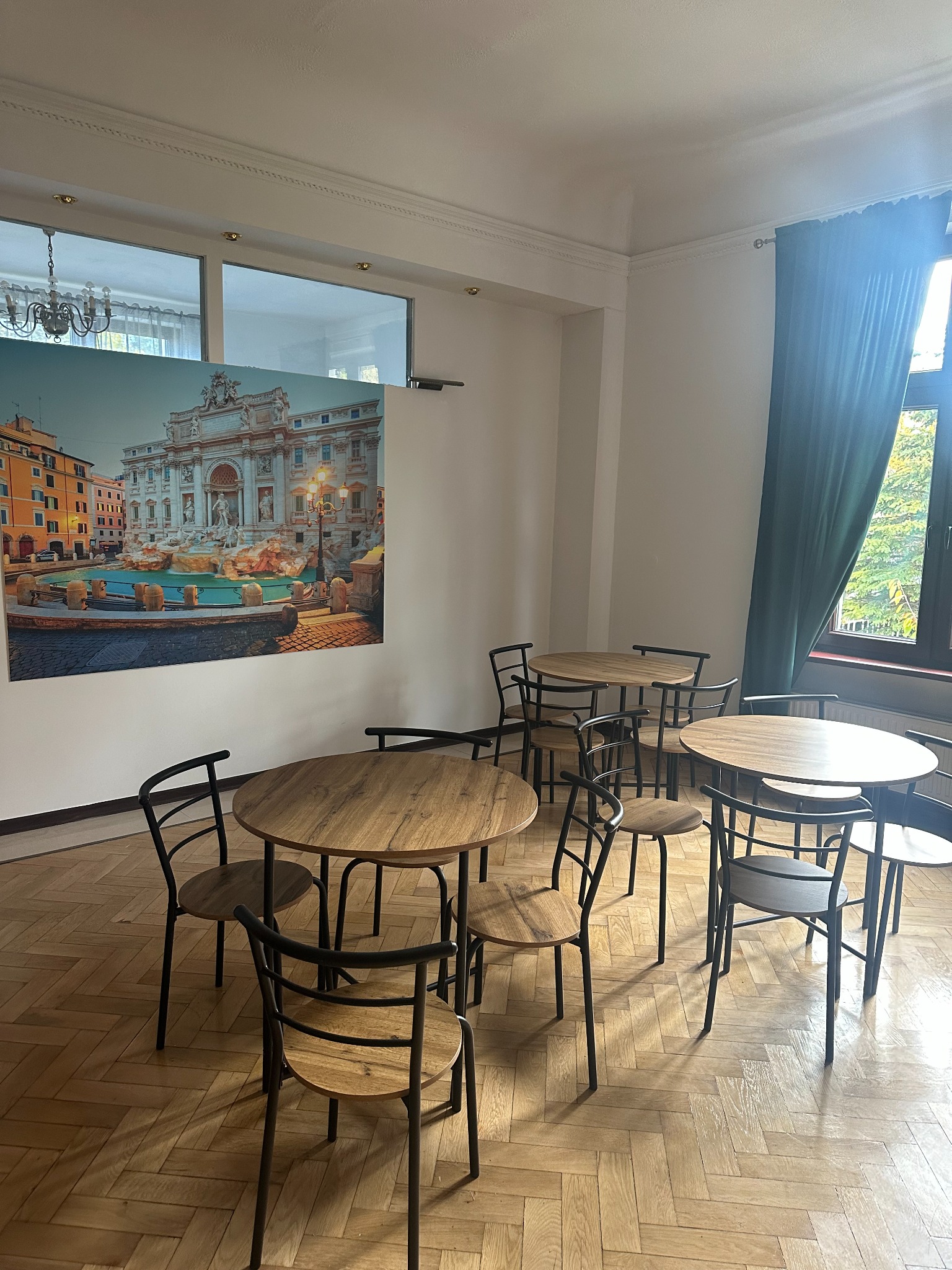 Sala kafeteryjna z drewnianymi stolikami i krzesłami na parkiecie w jodełkę, na ścianie widoczny plakat z Fontanną di Trevi.