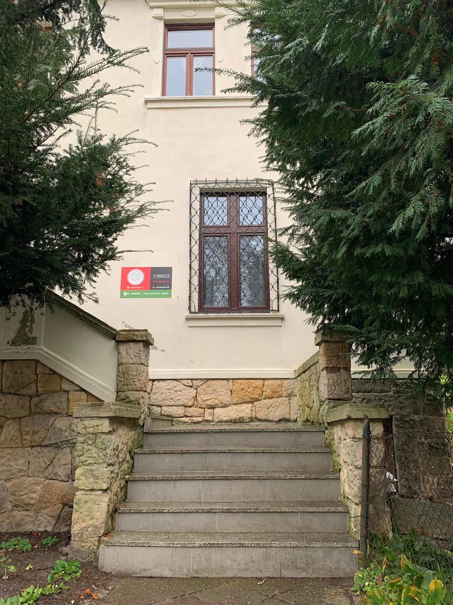 Wejście do budynku z kamiennymi schodami, otoczone zielonymi drzewami iglastymi, z tabliczką informacyjną wskazującą kierunek wejścia, sal lekcyjnych i biura.