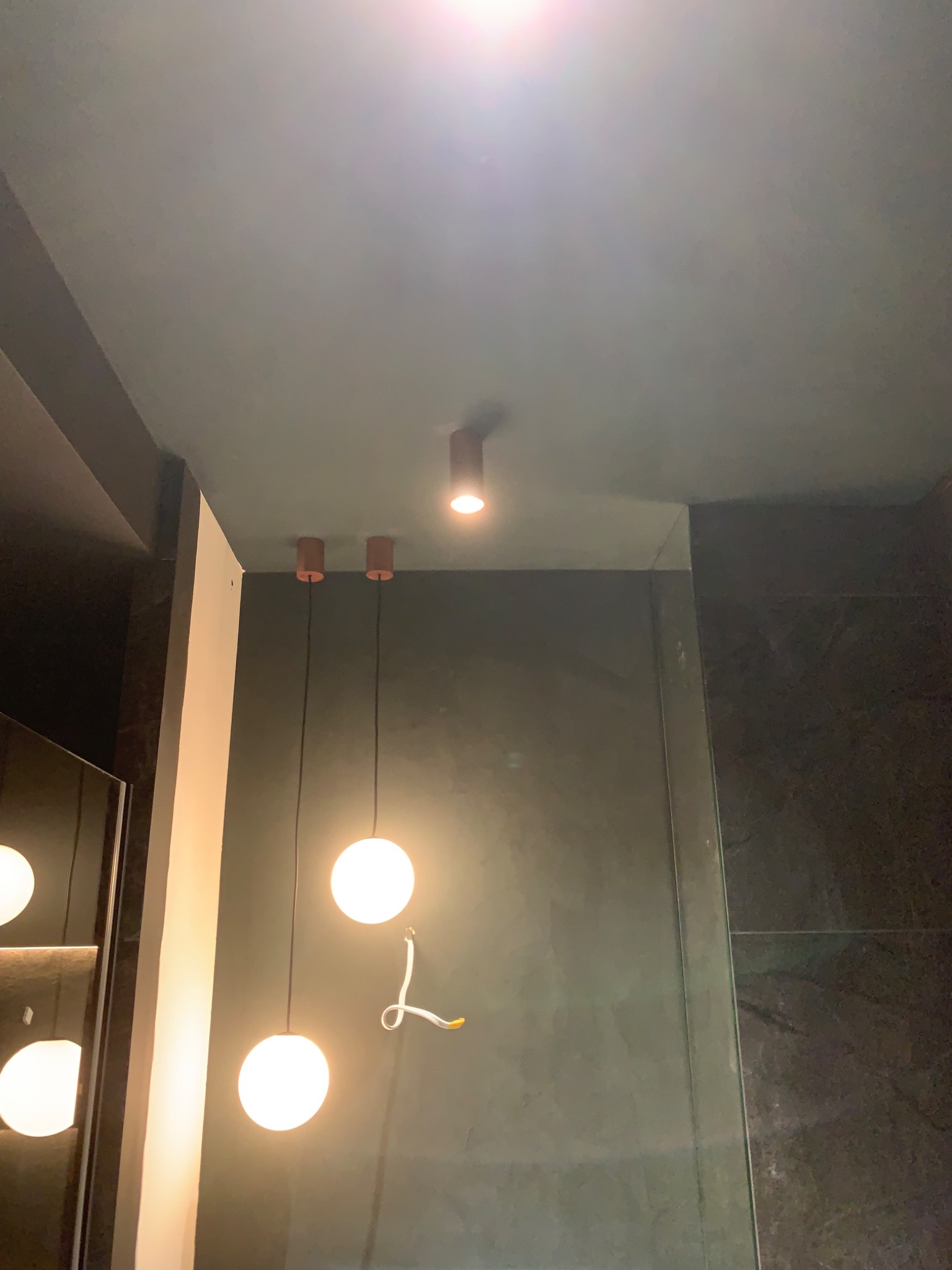 Zawieszone, kuliste lampy z przewodem w kolorze czarnym, zamontowane na suficie w łazience z ciemnymi, grafitowymi ścianami i szklaną kabiną prysznicową, widoczne przewody elektryczne.