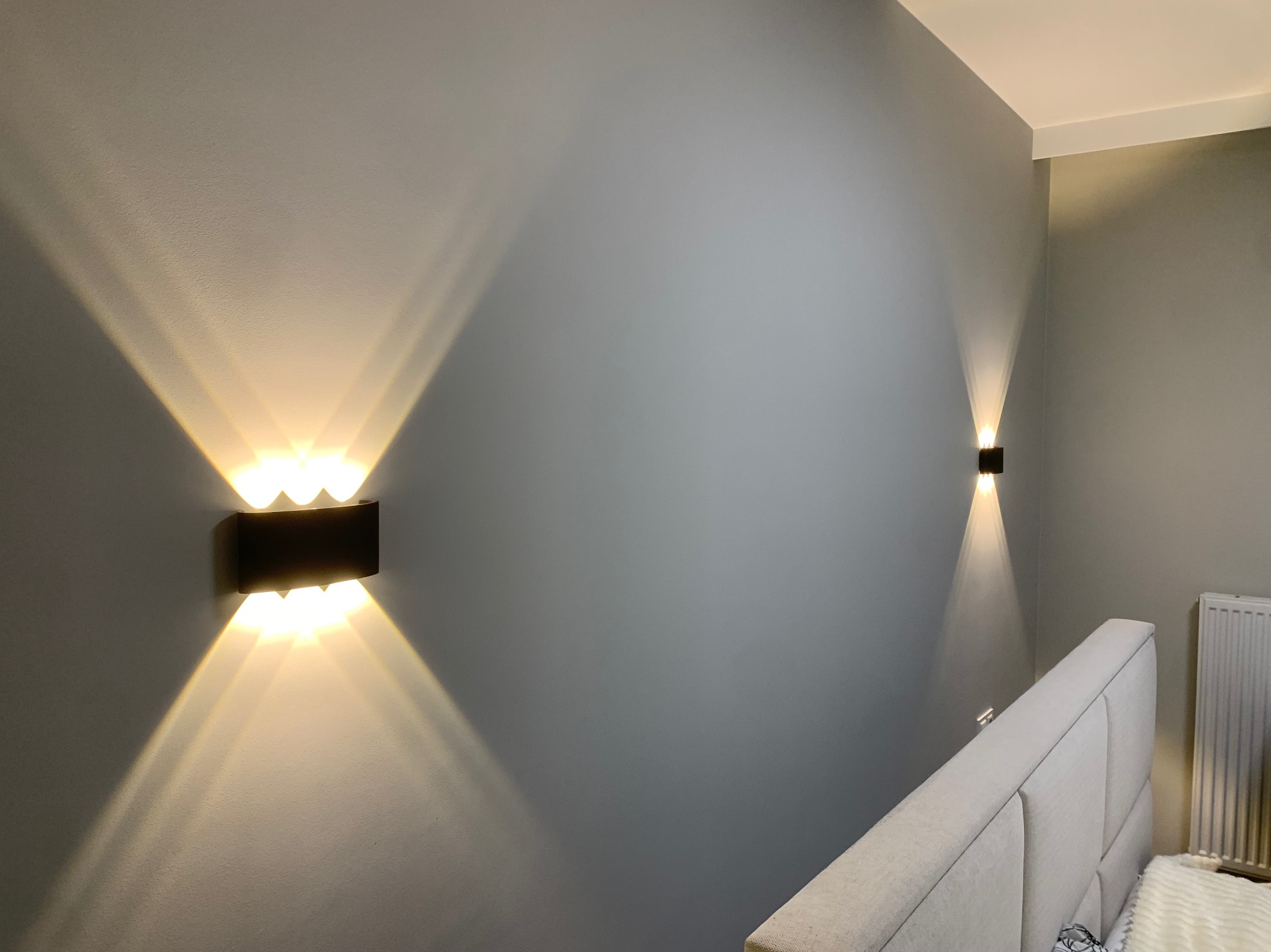 Dwa nowoczesne, czarne kinkiety ścienne z ciepłym, rozproszonym światłem, zamontowane na szarej ścianie w sypialni nad wezgłowiem łóżka.