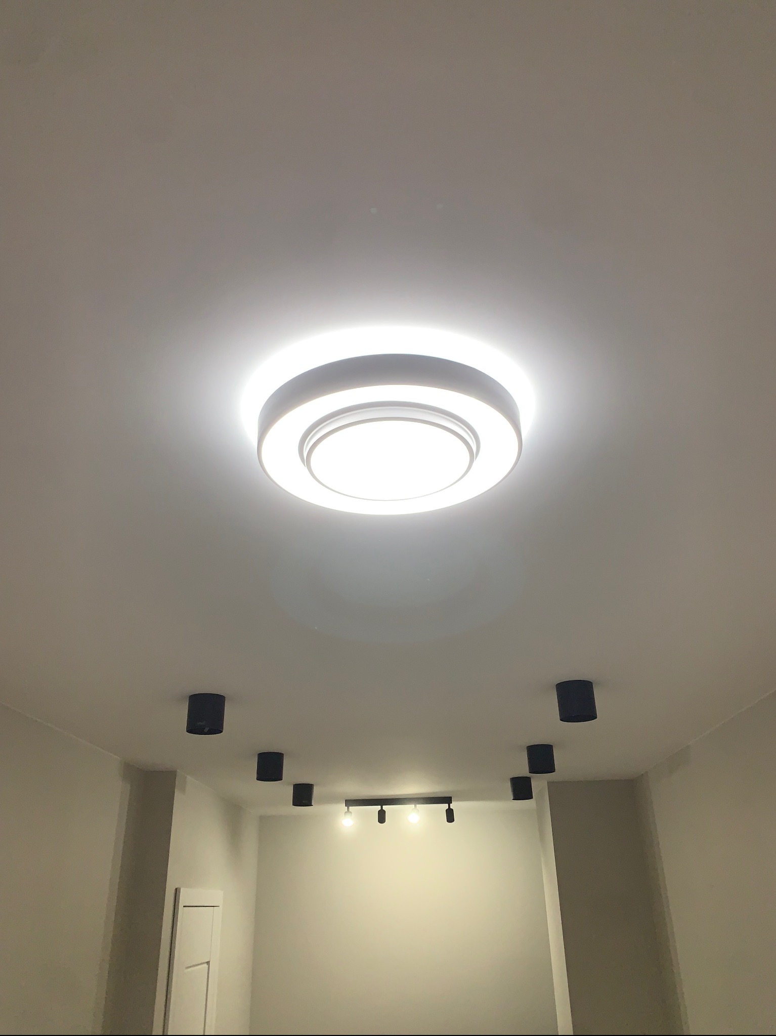 Centralnie umieszczona okrągła lampa sufitowa z trzema pierścieniami świetlnymi, otoczona rzędem czarnych, cylindrycznych reflektorów punktowych na białym suficie w nowo wykończonym wnętrzu.