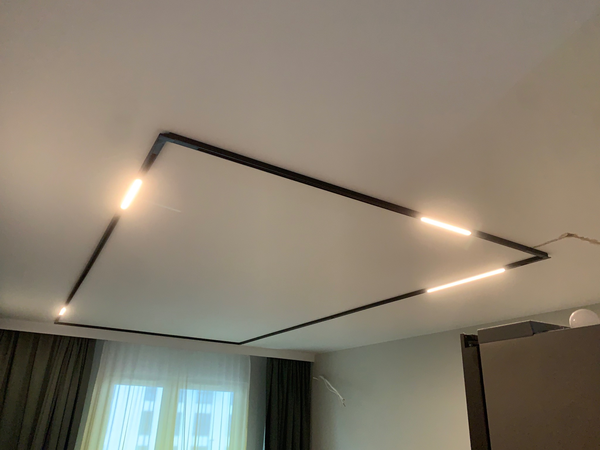 Nowoczesne oświetlenie sufitowe LED w kształcie prostokąta z czarną obudową, ciepłe światło, widok z dołu, fragment okna z zasłonami w tle.