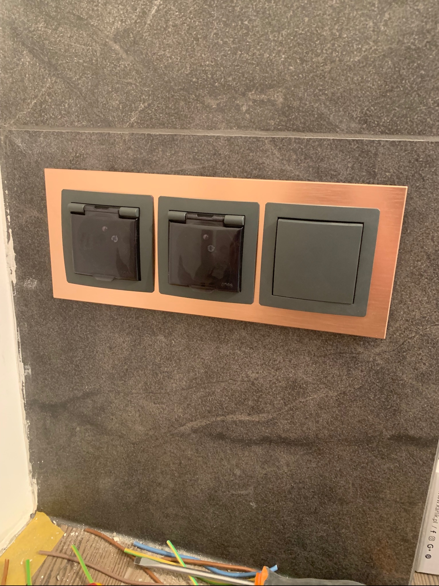Potrójne gniazdko elektryczne z klapkami ochronnymi i jednym standardowym włącznikiem, osadzone w miedzianej ramce na tle szarej ściany z imitacji kamienia. U dołu widoczne fragmenty przewodów...