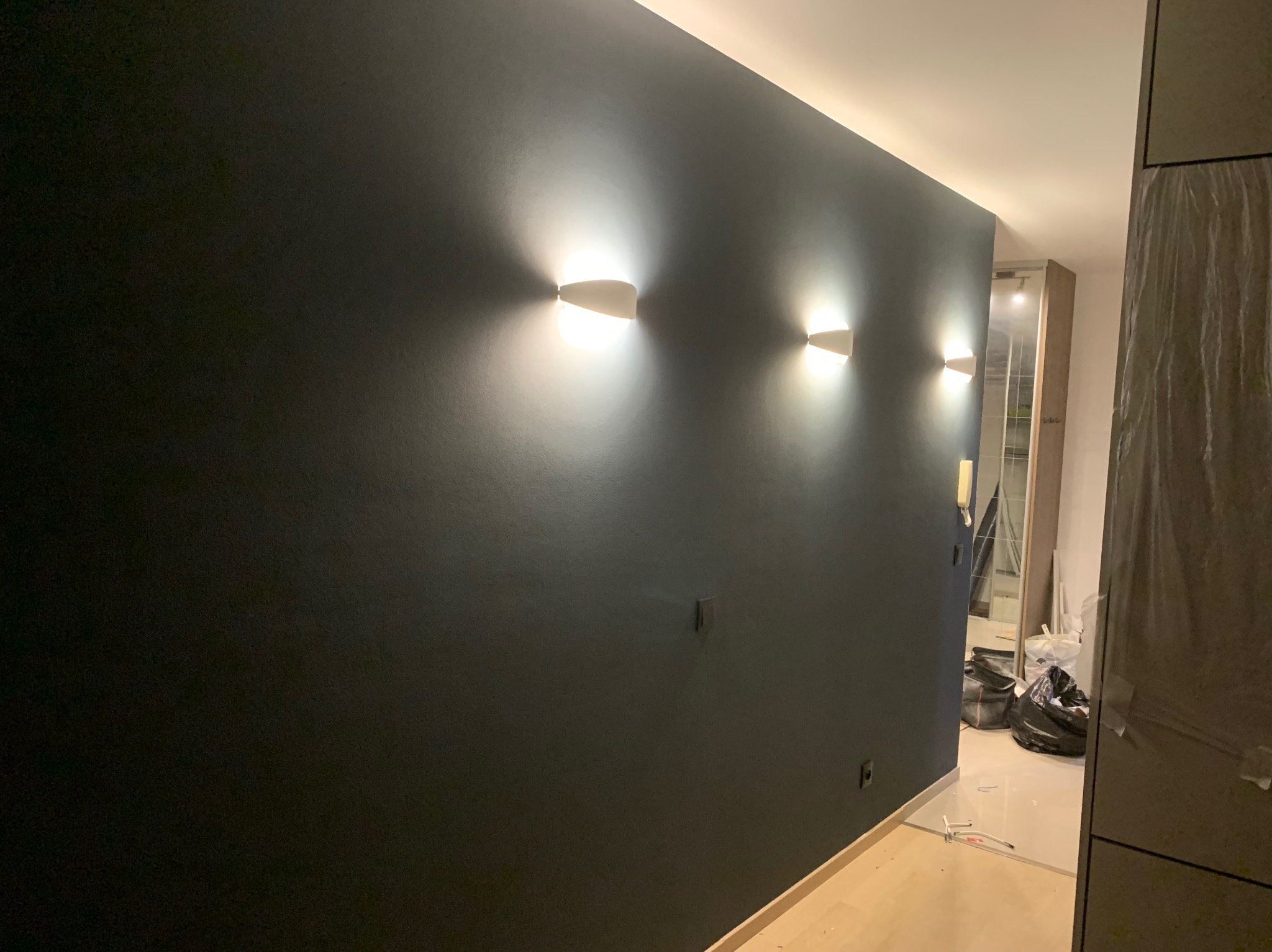 Ciemna ściana z trzema symetrycznie rozmieszczonymi, nowoczesnymi lampami ściennymi, dającymi ciepłe światło, widoczne gniazdka elektryczne i fragment pomieszczenia w trakcie wykończenia.