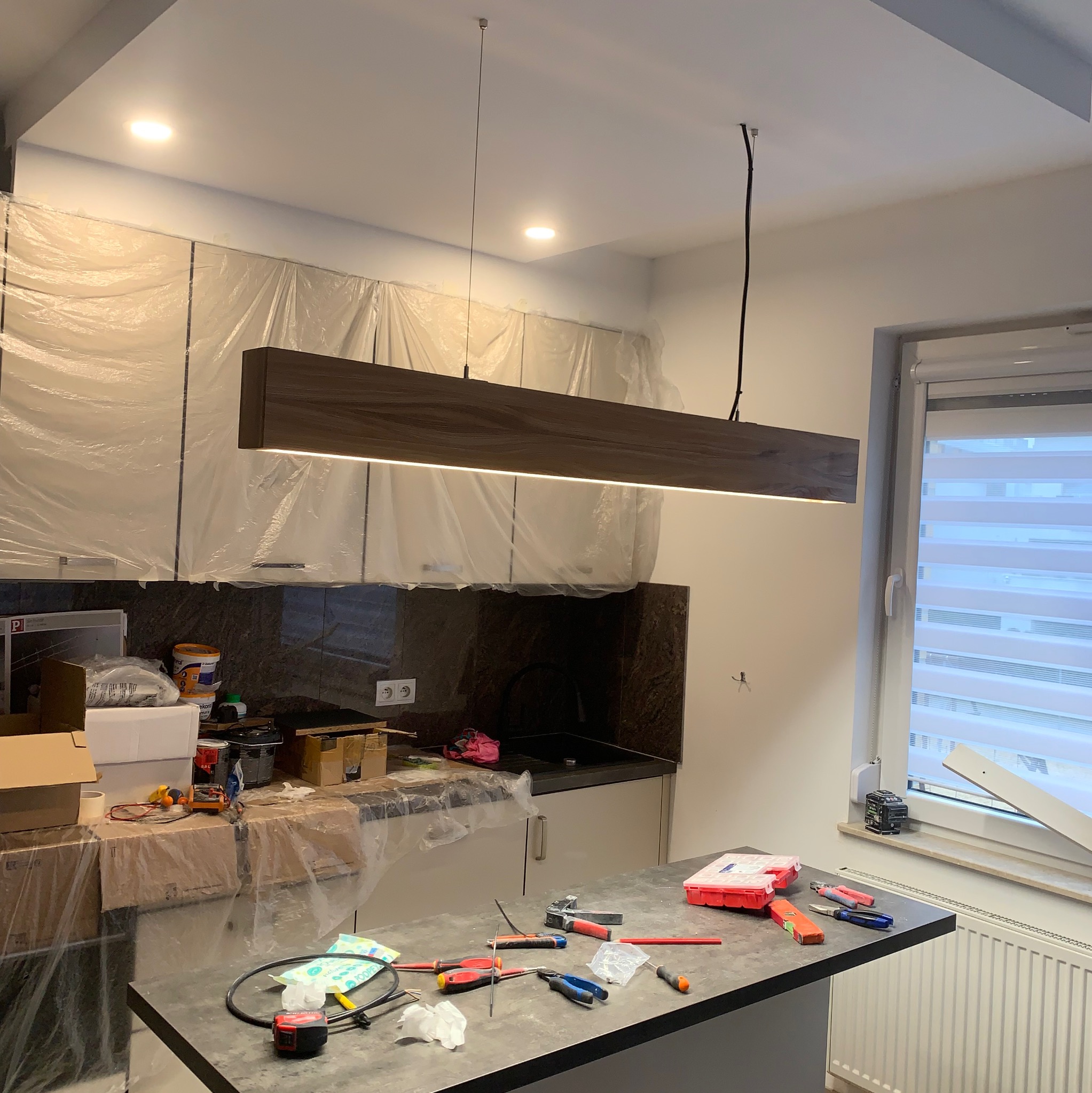 Nowoczesna, prostokątna lampa wisząca z drewnianym wykończeniem i wbudowanym oświetleniem LED, zamontowana w kuchni w trakcie remontu, z folią ochronną na szafkach i narzędziami na blacie.