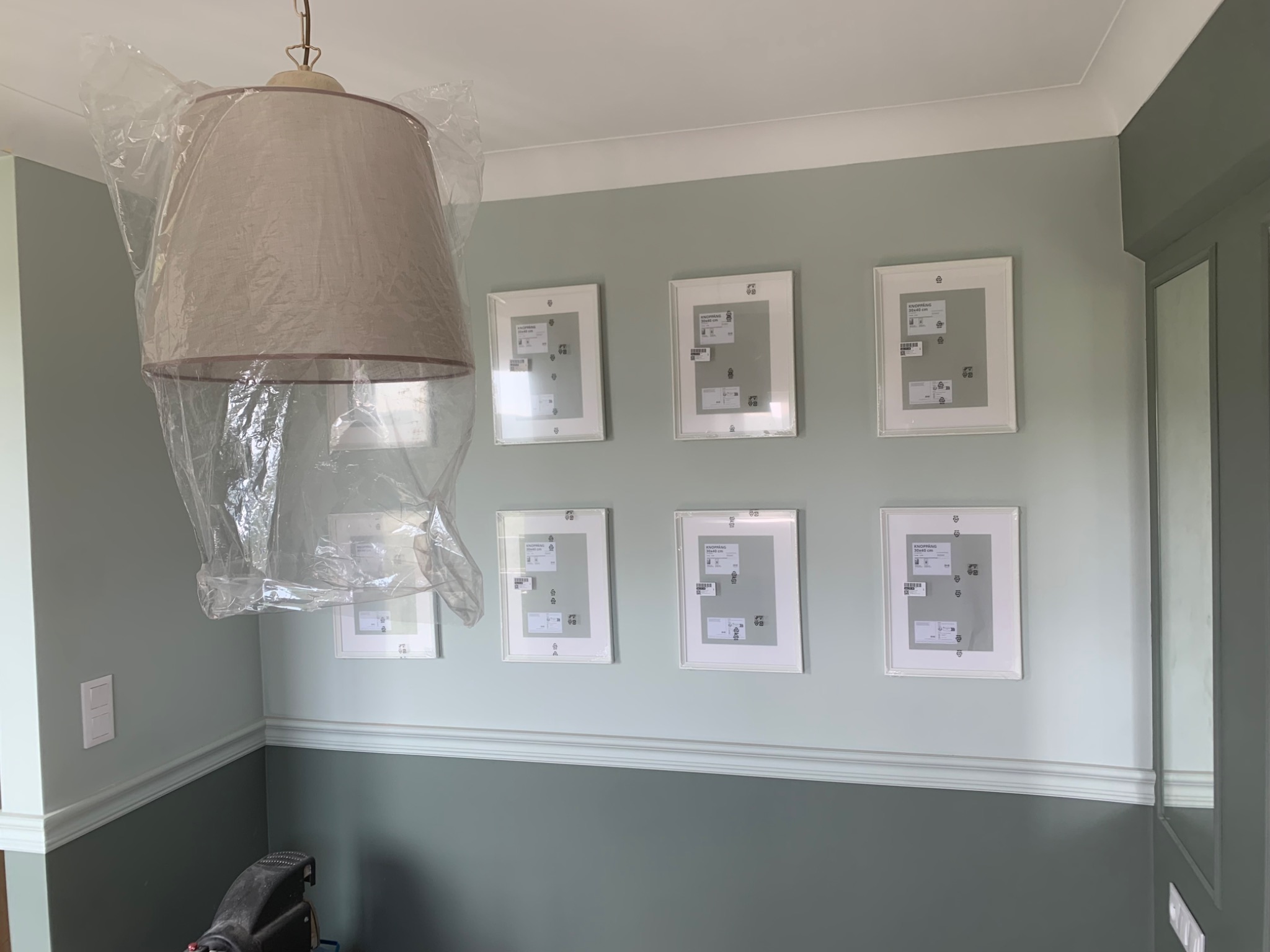 Lampa wisząca pod folią ochronną podczas montażu ramek na zdjęcia na ścianie w dwukolorowym pokoju. Widoczny fragment narzędzia.