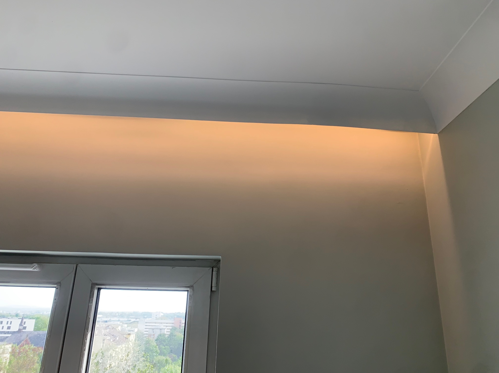 Ukryte oświetlenie LED w listwie przysufitowej, ciepłe światło odbija się od ściany. Okno z widokiem na miasto w tle. Nowoczesne rozwiązanie oświetleniowe.