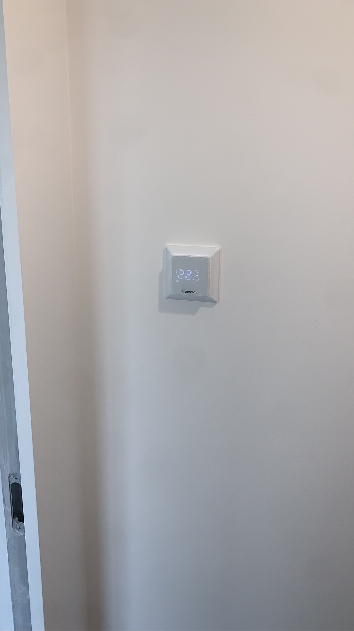 Biały, kwadratowy termostat pokojowy marki Termofol zamontowany na jasnej ścianie, wyświetlający temperaturę 22.3°C. Widoczny fragment ościeżnicy po lewej.