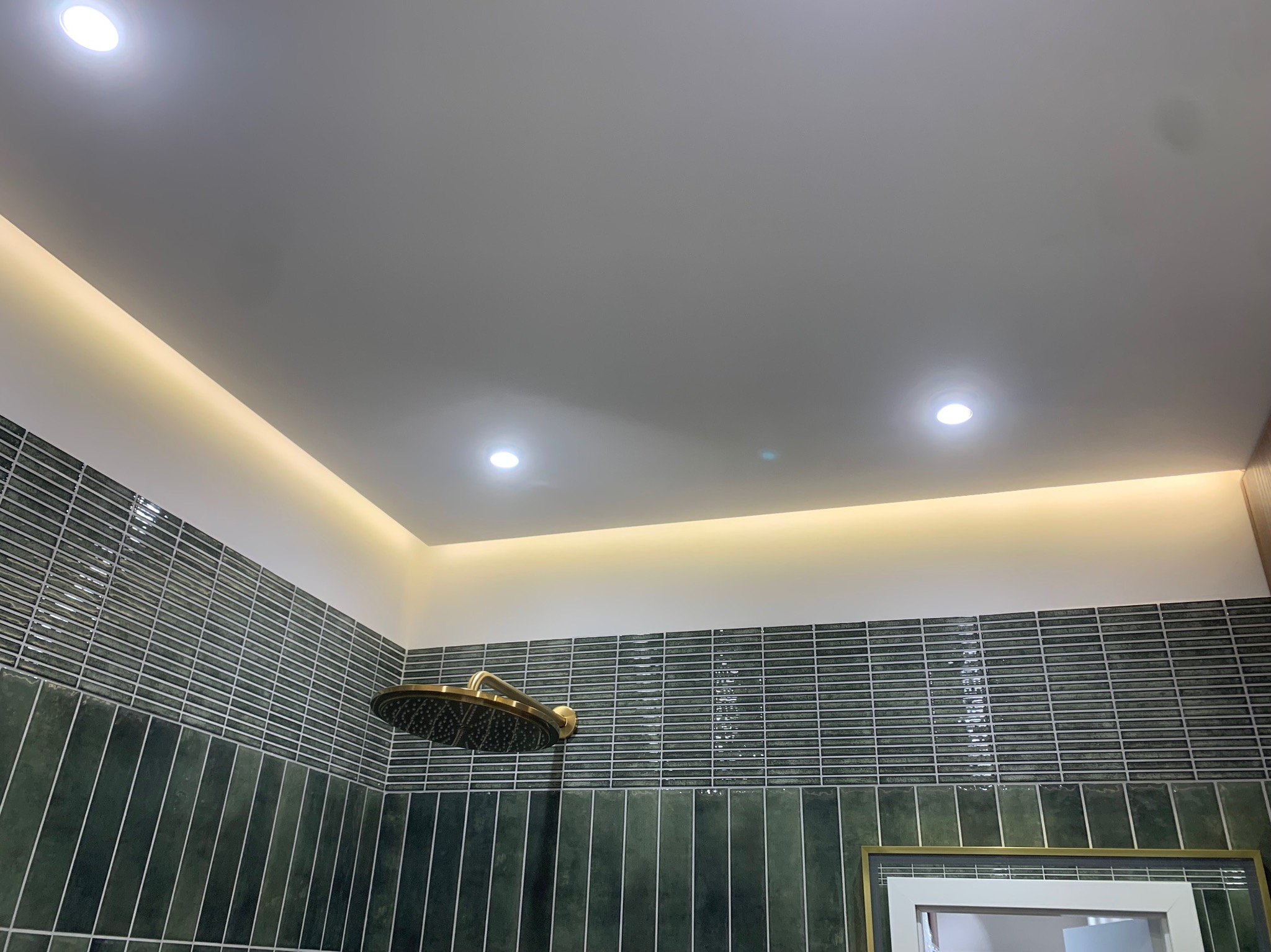 Nowoczesna łazienka z zielonymi płytkami, złotą deszczownicą i podświetleniem LED na suficie. Minimalistyczny design i eleganckie wykończenie.