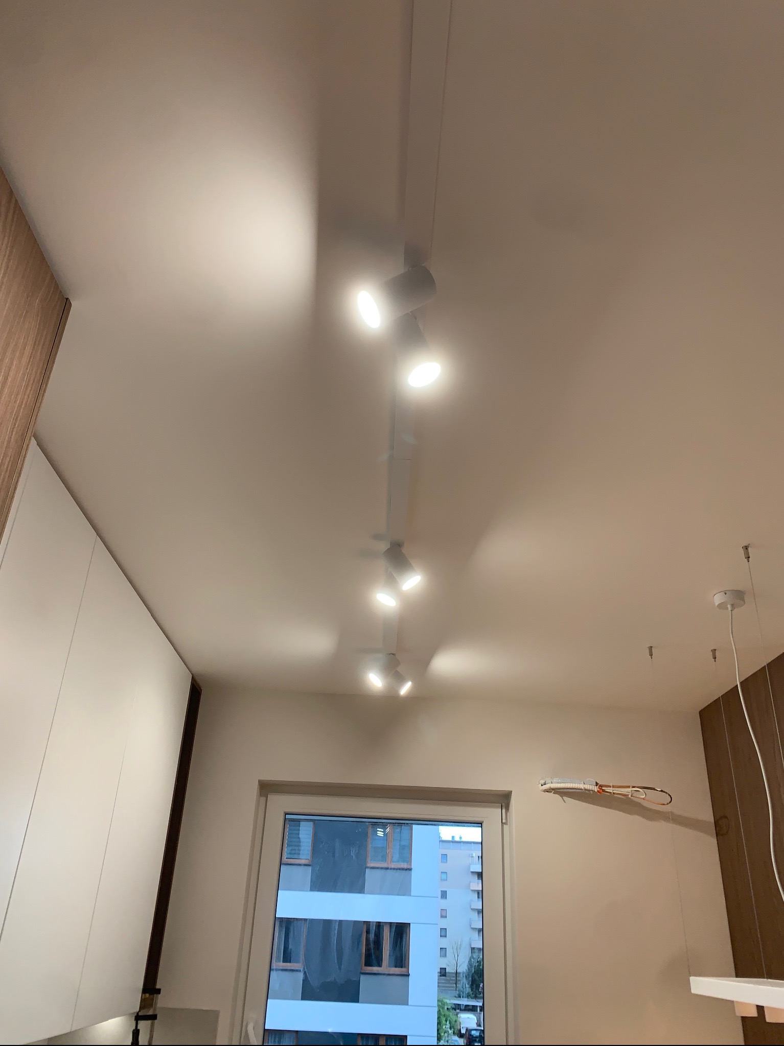 Szyna z reflektorami sufitowymi w nowoczesnym wnętrzu. Widoczne okno i fragmenty zabudowy kuchennej. Reflektory oświetlają pomieszczenie.