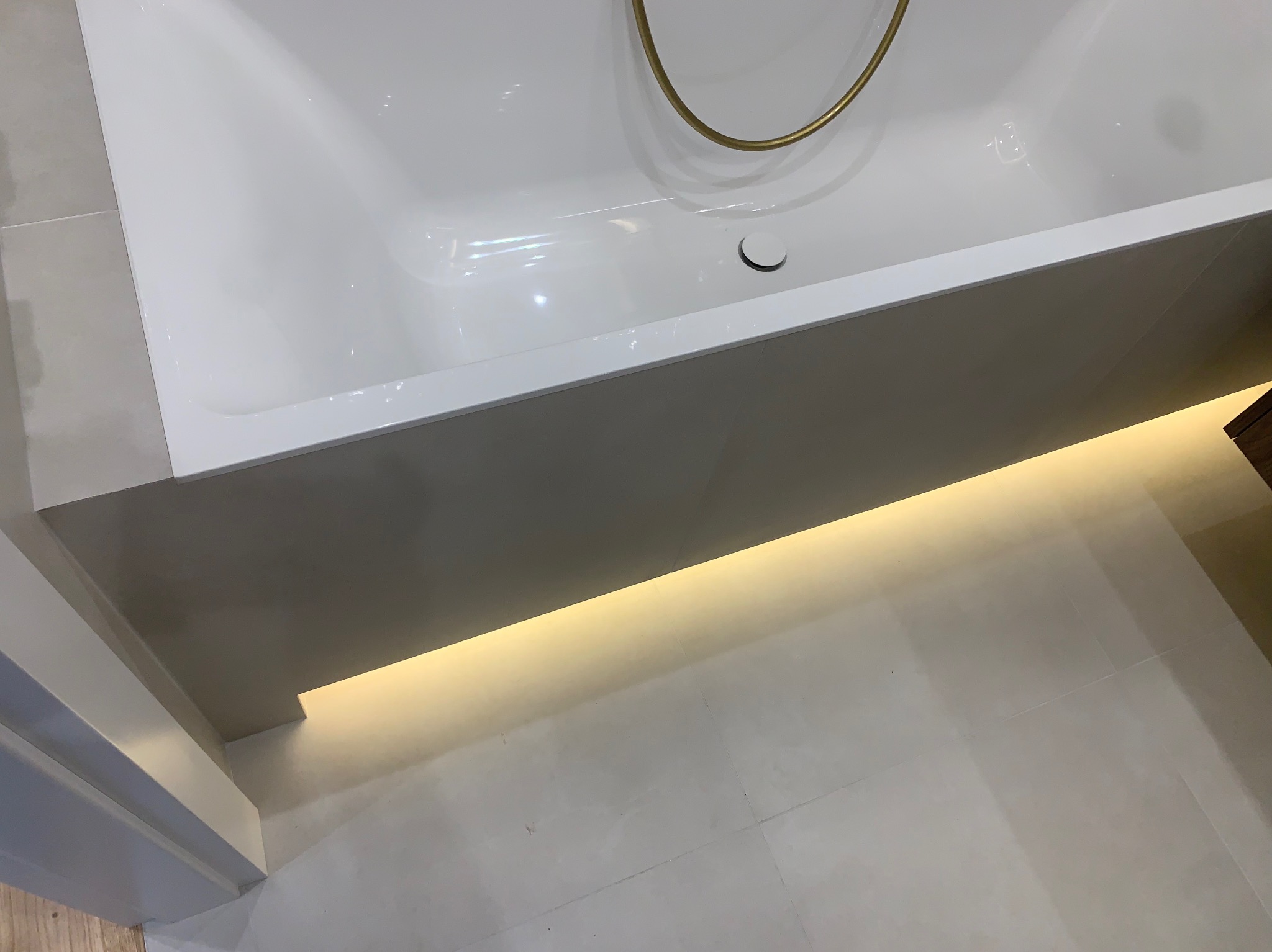 Nowoczesna, biała wanna z podświetleniem LED w obudowie. Widoczna złota armatura i jasne płytki podłogowe. Minimalistyczny design łazienki.