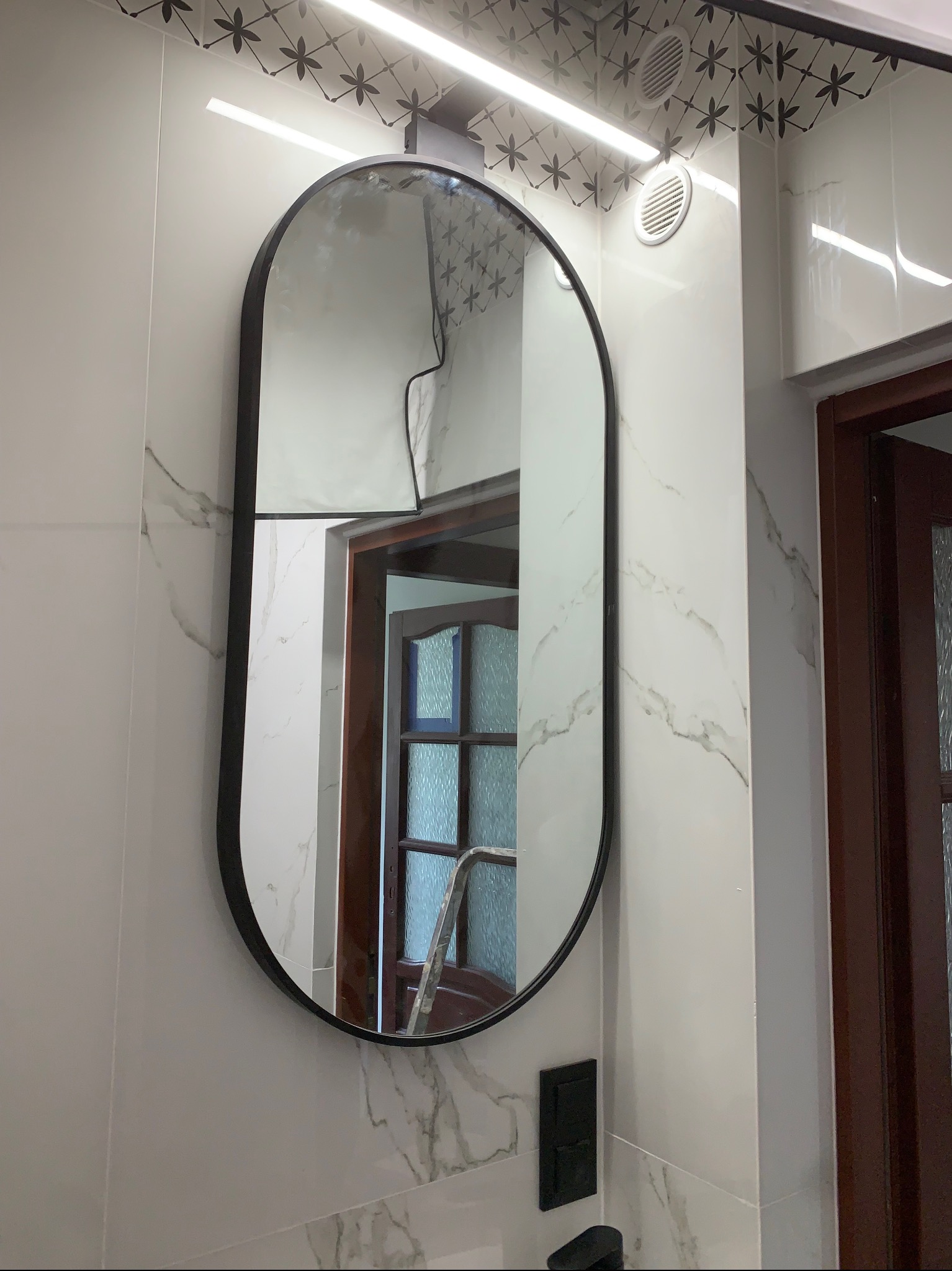 Oświetlenie LED nad owalnym lustrem w łazience z marmurowymi płytkami i czarnym gniazdkiem. Widoczny fragment kratki wentylacyjnej i drzwi w odbiciu lustra.