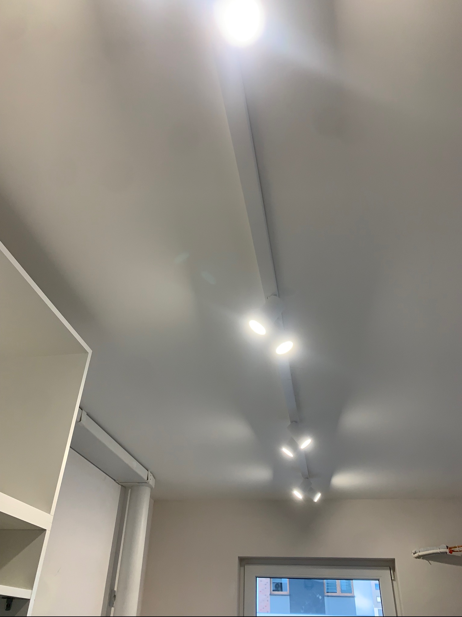 Biały sufit z zamontowaną szyną oświetleniową i reflektorami punktowymi LED, tworzącymi jasne, równomierne światło. Widok z dołu, fragment mebla w lewym dolnym rogu.