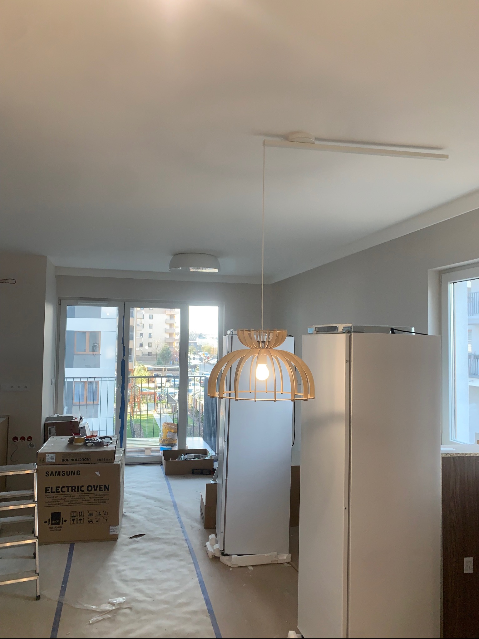 Nowoczesna lampa wisząca z drewnianymi elementami w jasnym wnętrzu w trakcie wykończenia. Widoczne kartony po sprzęcie AGD i zabezpieczona podłoga.