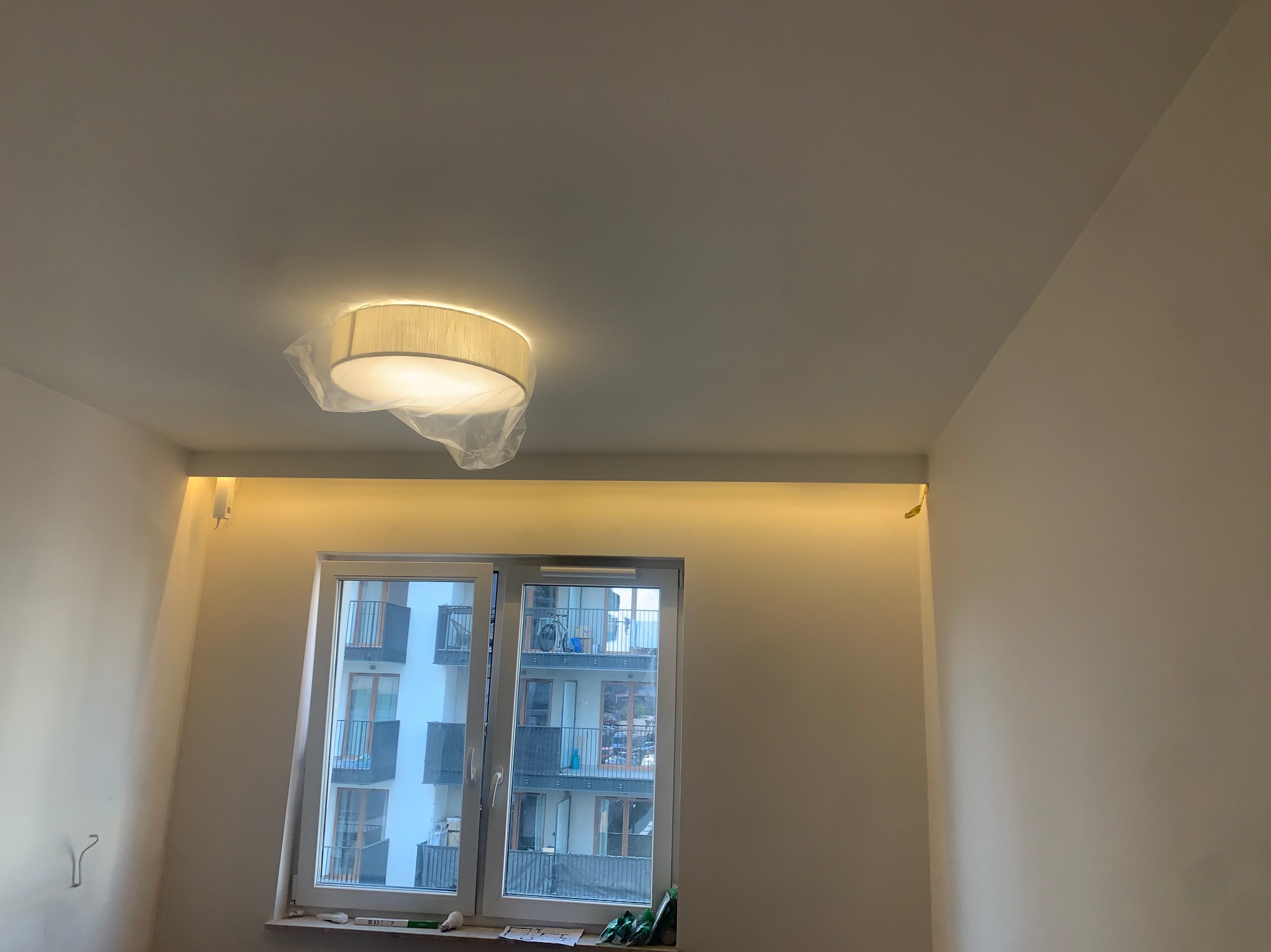 Nowoczesny pokój z oknem: zamontowane oświetlenie sufitowe w folii ochronnej i listwa LED nad oknem. Widok na budynek z balkonami przez okno.