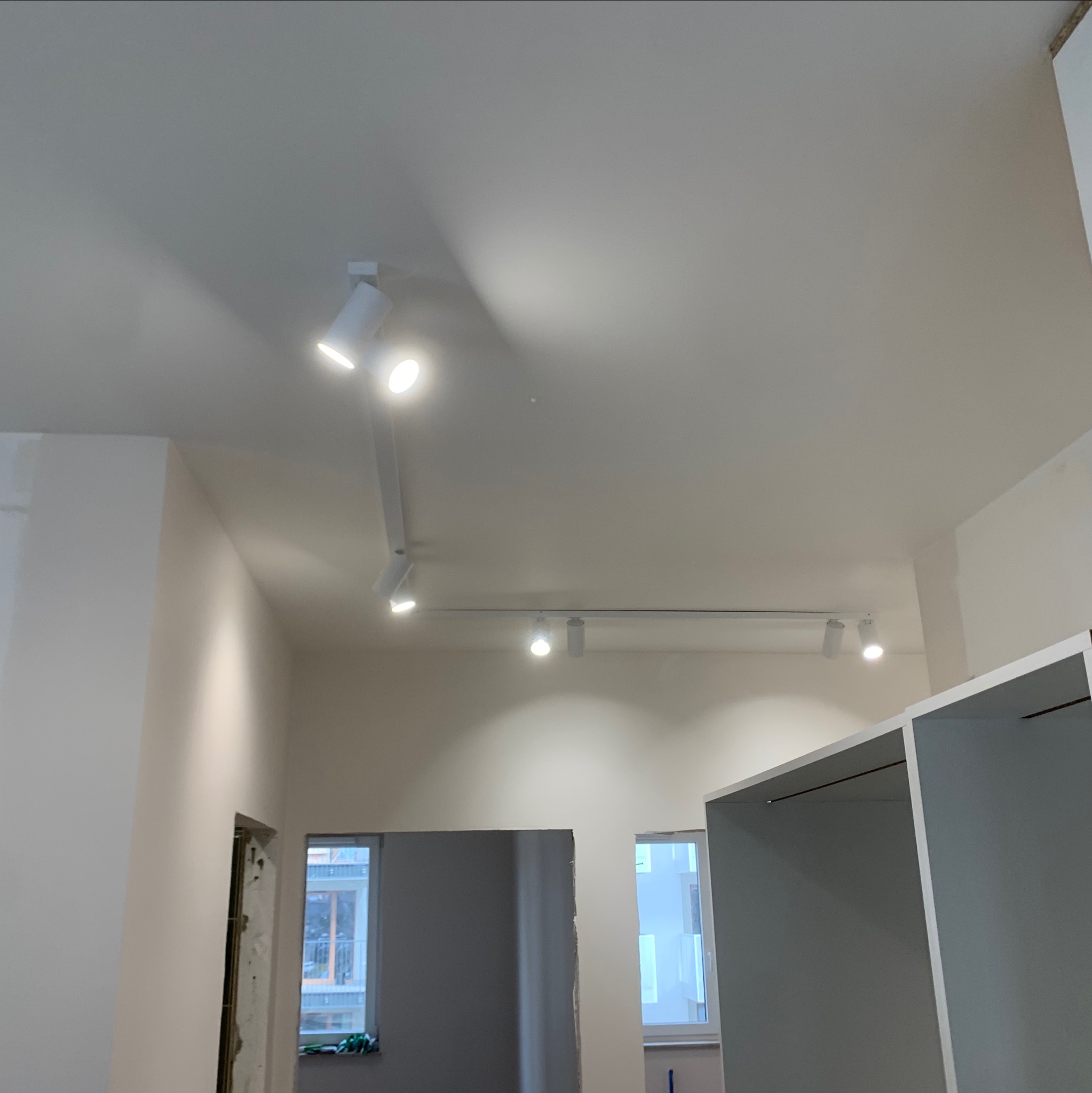 Białe reflektory szynowe zamontowane na suficie w jasnym wnętrzu. Widoczne okna i fragmenty mebli. Oświetlenie punktowe, nowoczesny design.