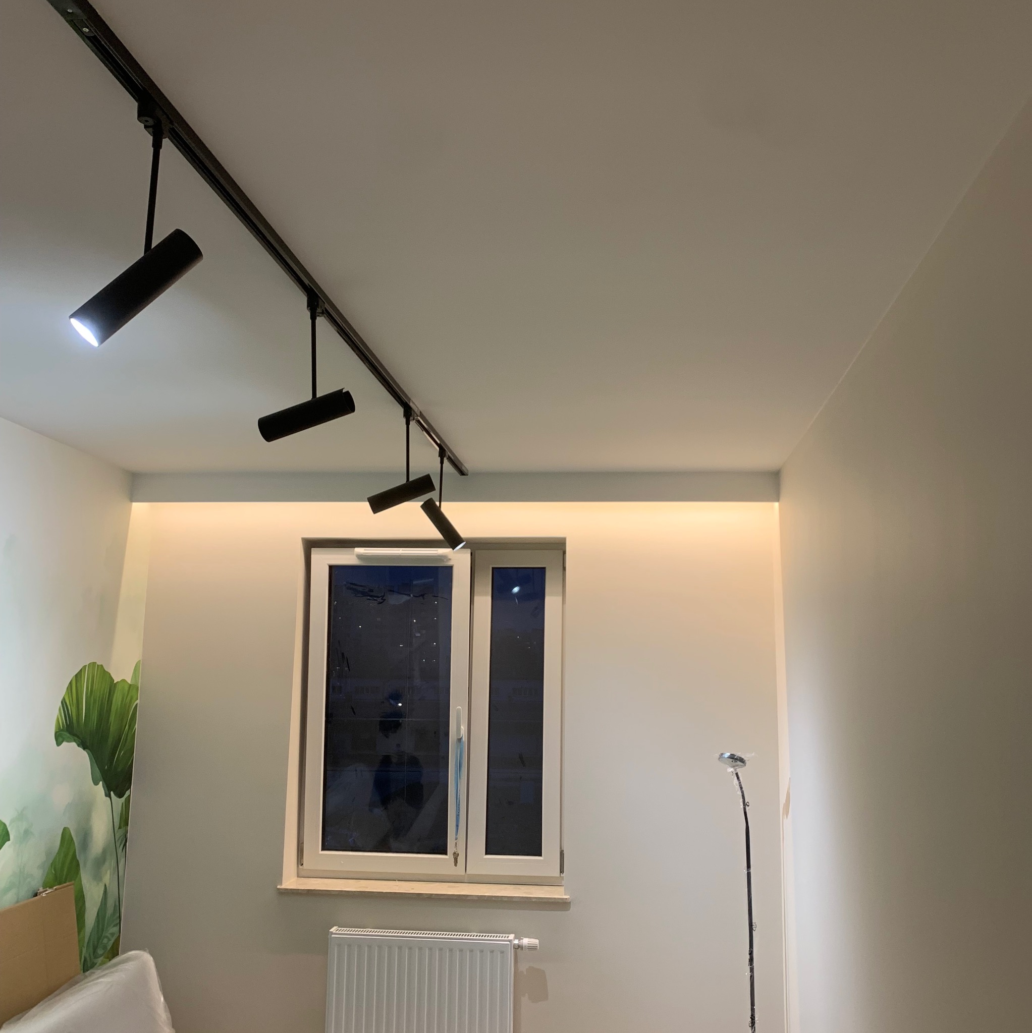 Nowoczesne oświetlenie szynowe z czarnymi reflektorami w jasnym wnętrzu, dodatkowo listwa LED nad oknem. Widoczny fragment tapety z motywem roślinnym.