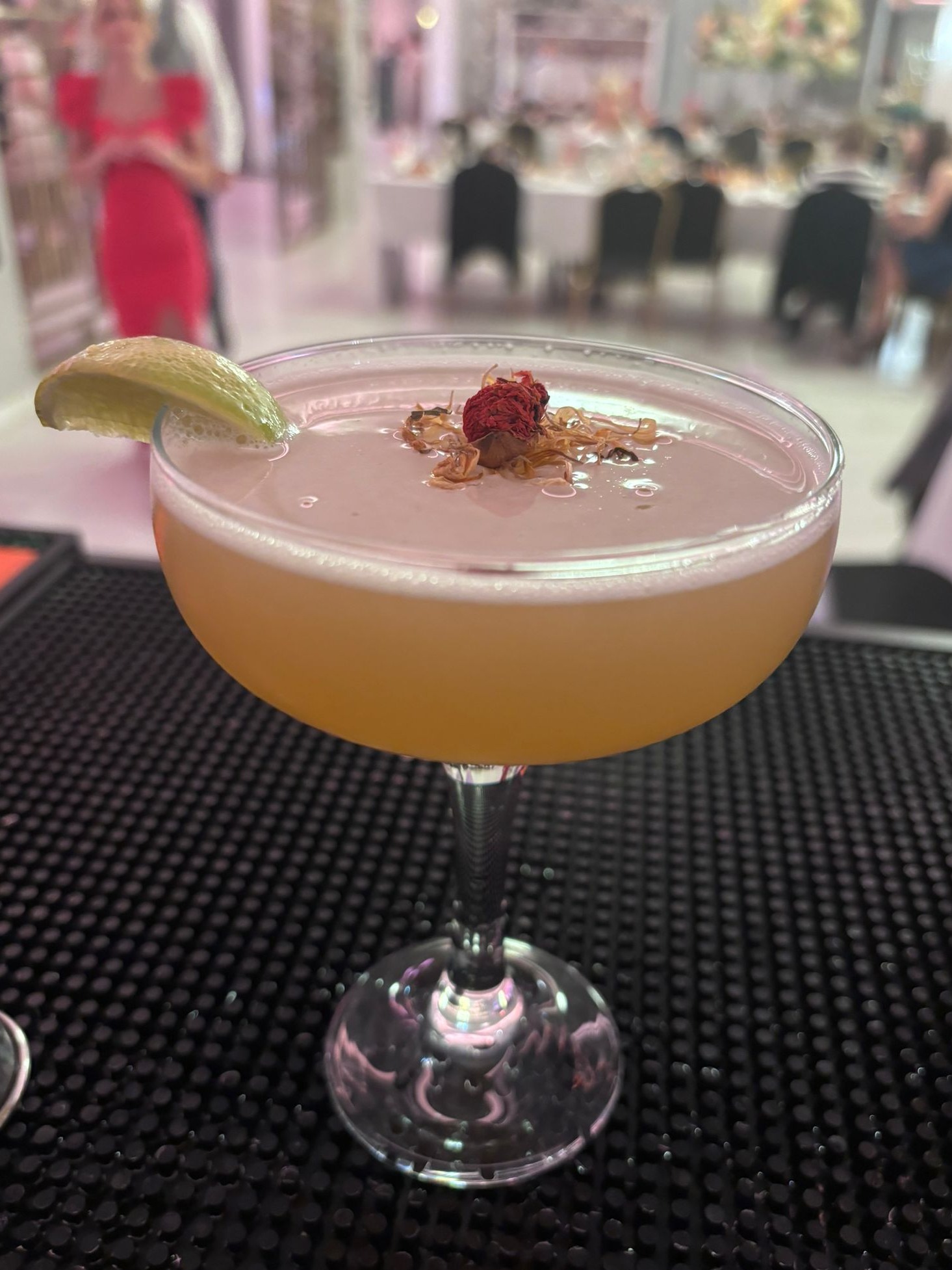 Elegancki koktajl w kieliszku typu coupe, ozdobiony suszoną różą i skórką limonki, ustawiony na czarnej macie barmańskiej, z rozmytym tłem sali bankietowej.