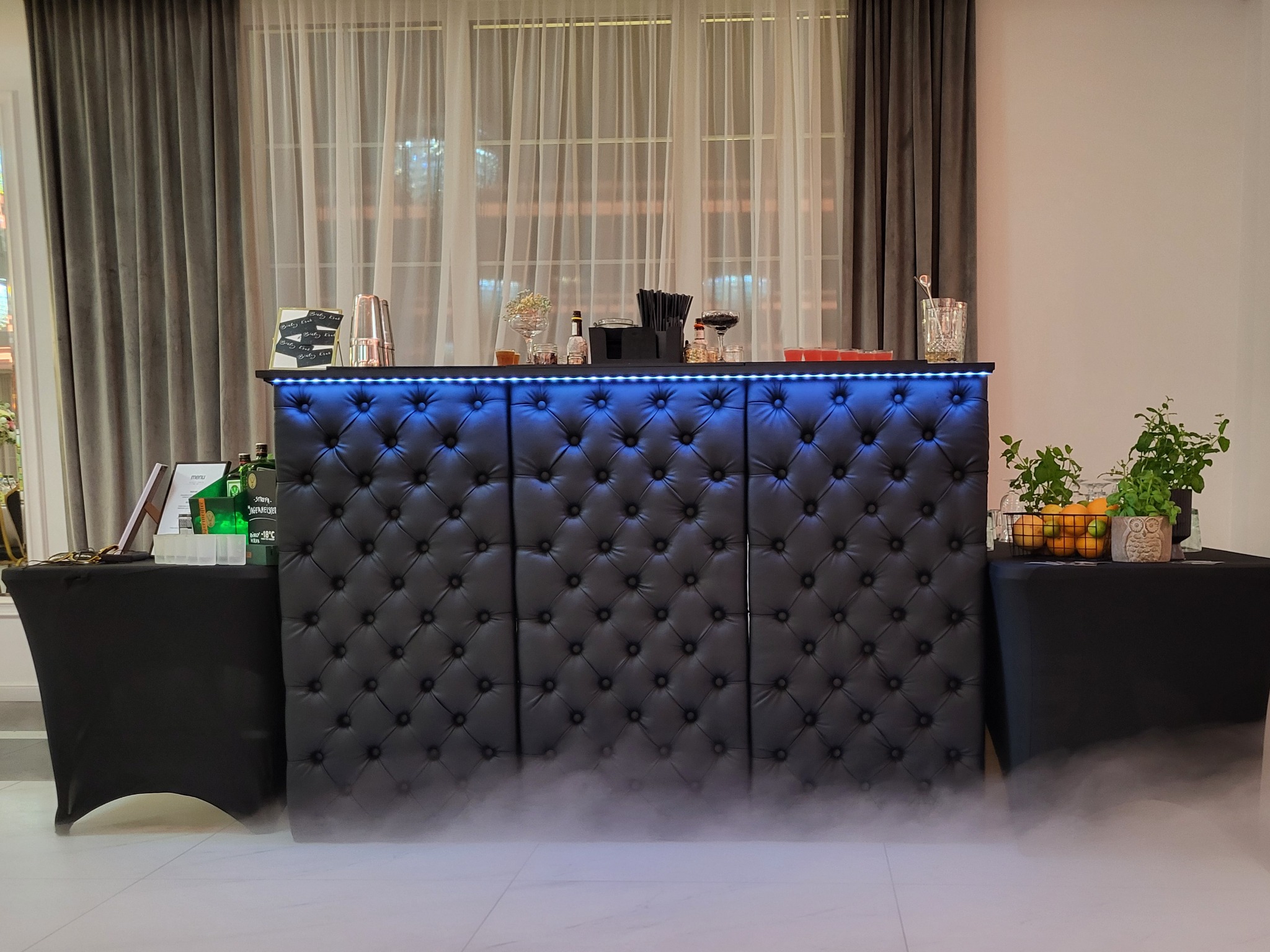 Elegancki bar z pikowaną, czarną skórą, podświetlony niebieskim światłem LED, z akcesoriami barmańskimi i dekoracjami, otoczony lekką mgłą.