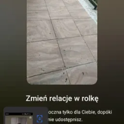 Balkon z brązowymi płytkami podłogowymi w trakcie układania, widoczne krzyżyki dystansowe, czarna metalowa balustrada z ozdobnymi elementami.