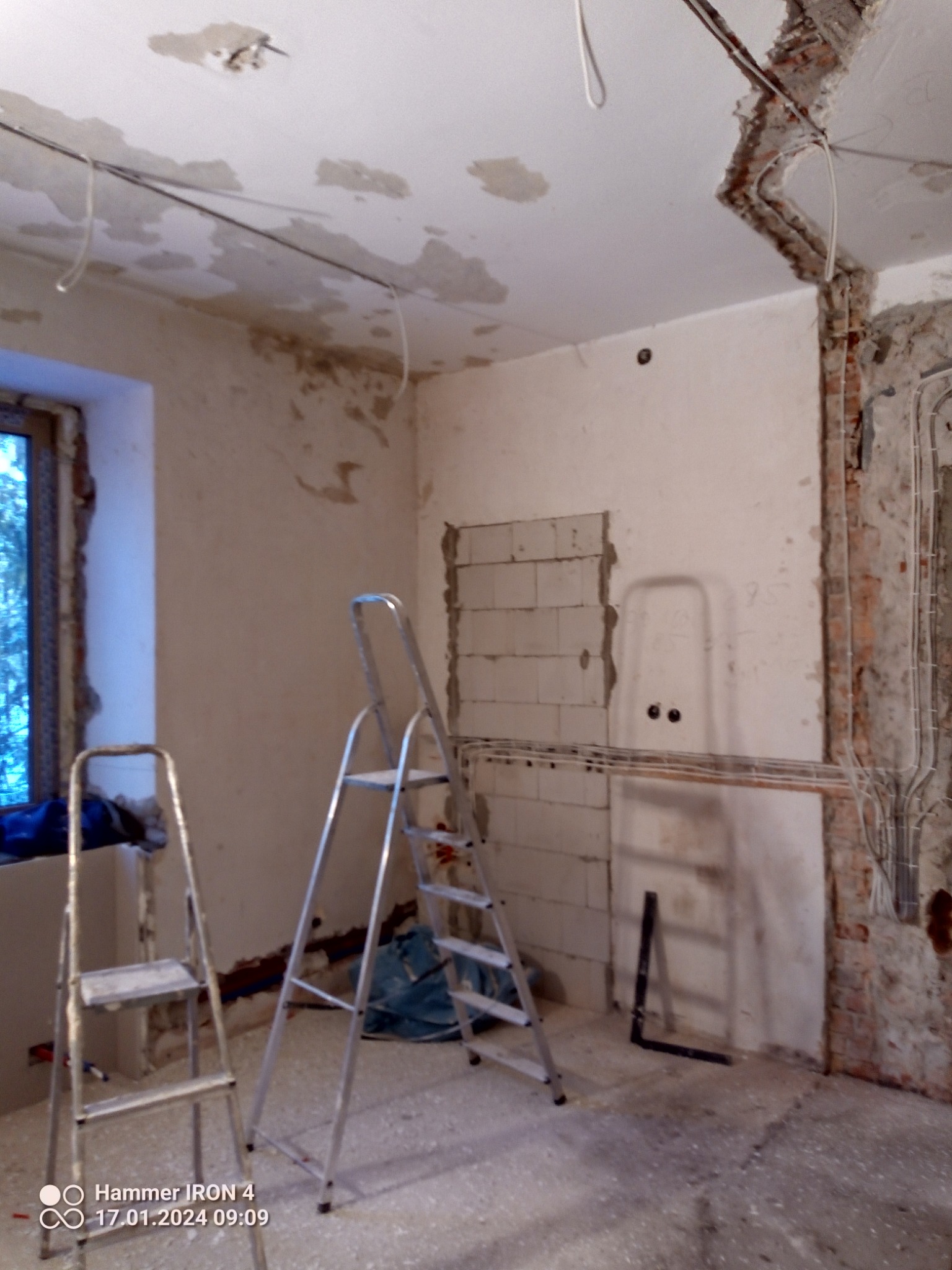Pokój w trakcie remontu: odsłonięta cegła, instalacja elektryczna, ślady po skuciu tynku, dwie drabiny aluminiowe, widoczne przewody elektryczne na suficie.