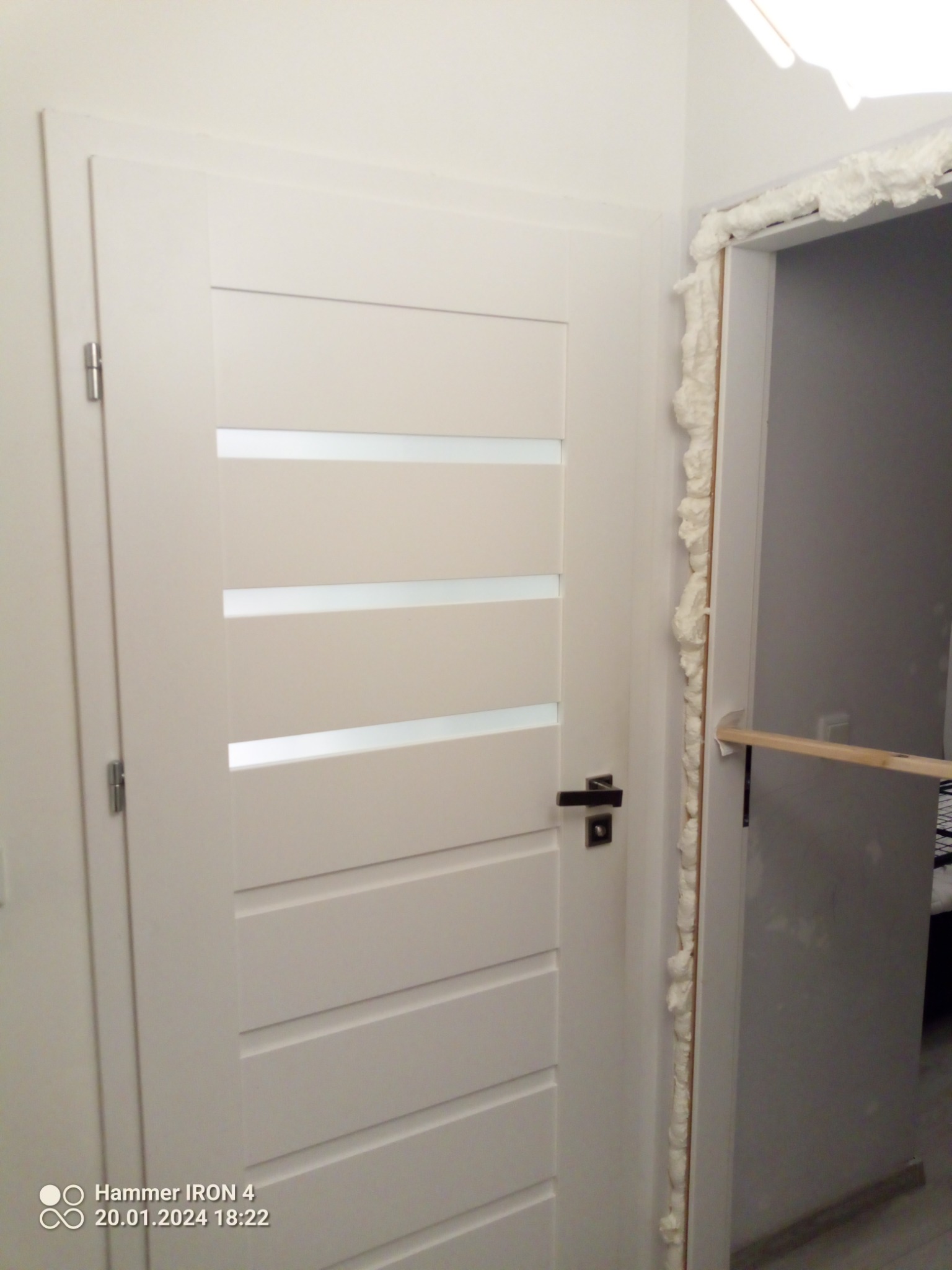 Białe drzwi wewnętrzne z poziomymi przeszkleniami, osadzone w ościeżnicy wypełnionej pianką montażową, widoczne ślady niedokończonego montażu.