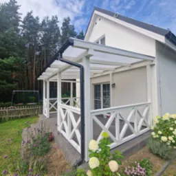 Biała pergola z przezroczystym dachem przylegająca do jasnego domu, z dekoracyjną balustradą i widokiem na ogród z kwiatami.