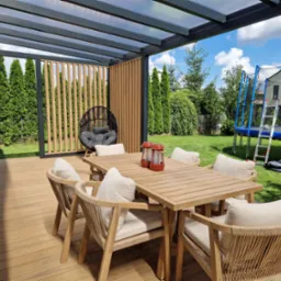 Zadaszony taras z drewnianą podłogą, stołem i krzesłami w stylu boho. W tle zielony ogr&oacute;d z trampoliną i tujami. Widoczna ażurowa ścianka i wiszące krzesło.