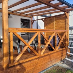 Drew-Mar-Styl Marcin Włodarczak - Zadaszony taras drewniany z balustradą w kształcie krzyży, z widoczną osłoną grilla i drabinką opartą o bok tarasu.